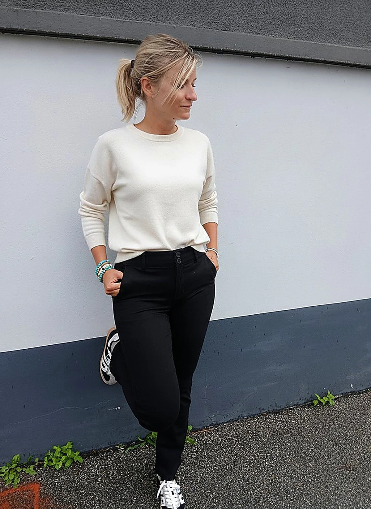 Laurie en pantalon Bibette noir, pull écru et baskets, adossée au mur de la boutique
