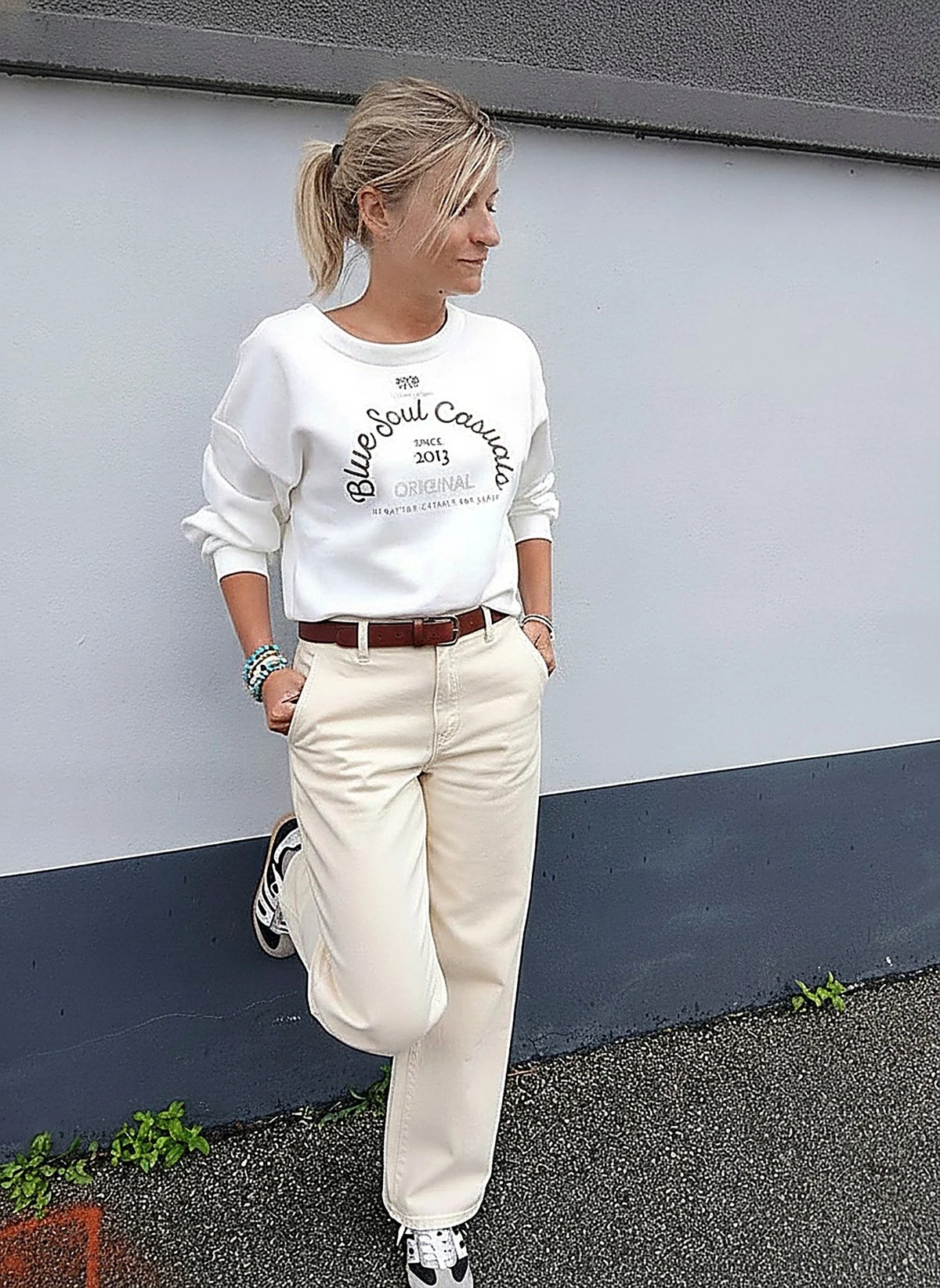 Laurie en pantalon Colette twill à ceinture écru, adossée au mur de la boutique