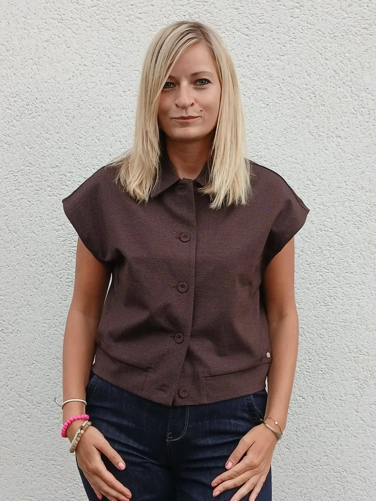 Laurie en chemise marron à manches courtes et jean brut, devant un mur blanc