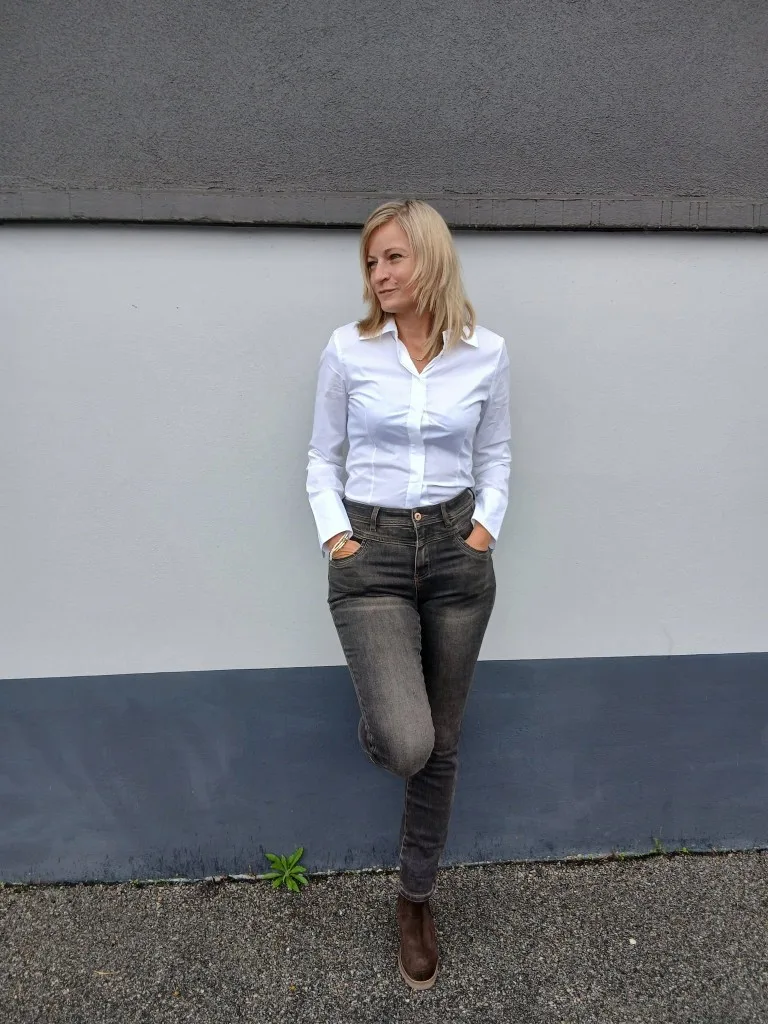 Laurie en chemise blanche et jean sombre, adossée au mur, une jambe croisée
