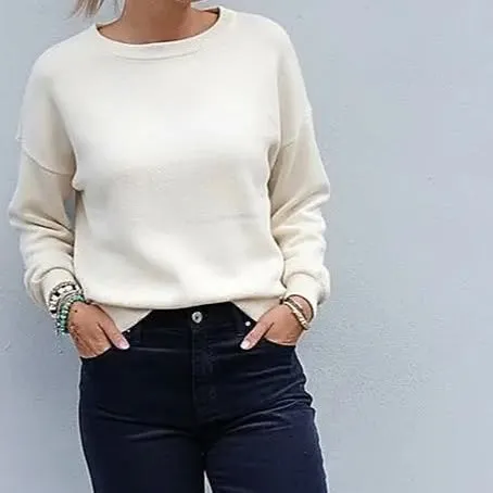 Pull crème à col rond et jean brut, porté contre un mur clair